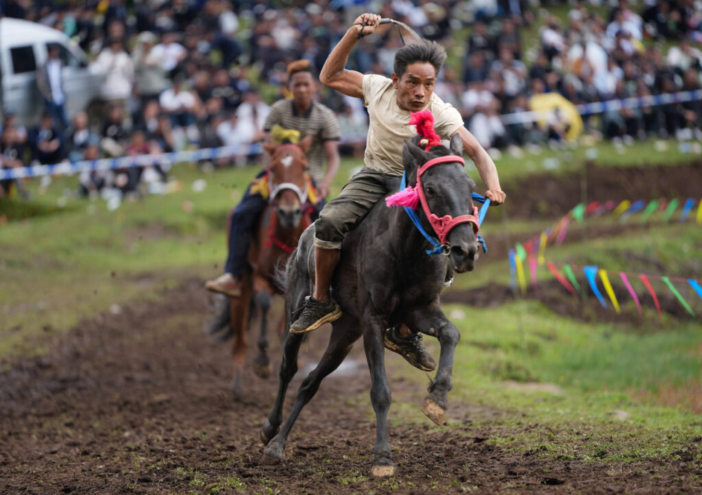 Yi man riding bareback on a galloping black horse during a traditional bareback horse race at Yi Torch Festival, with colorful flags and spectators in the background