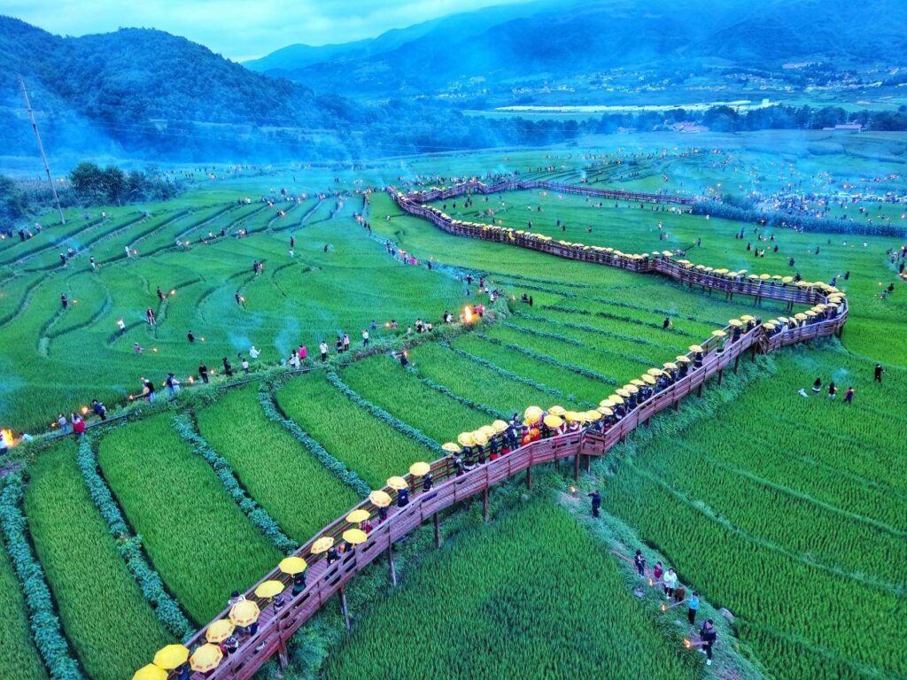 Aerial view of Yi Torch Festival procession along terraced rice paddies, visitors carrying yellow umbrellas on a wooden boardwalk with small torches lighting the path