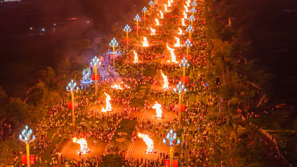 Aerial night view of Yi Torch Festival celebration with rows of large bonfires along a crowded plaza, decorated blue torch poles and mountains in the background